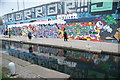 View of street art on the wall of a warehouse on the Hertford Union Canal towpath reflected in the canal