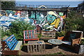 View of a wall of street art, benches made of wooden palettes and a table made from a cable reel in the Nomadic Community Garden